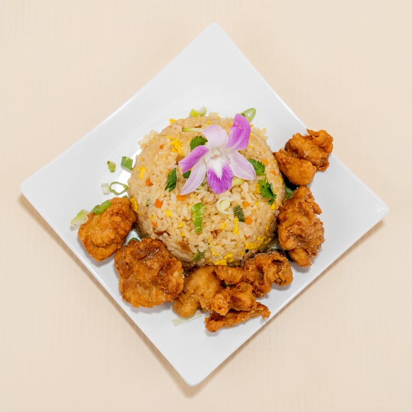 Fried rice with vegetables and a purple orchid, surrounded by crispy fried chicken pieces on a white plate.