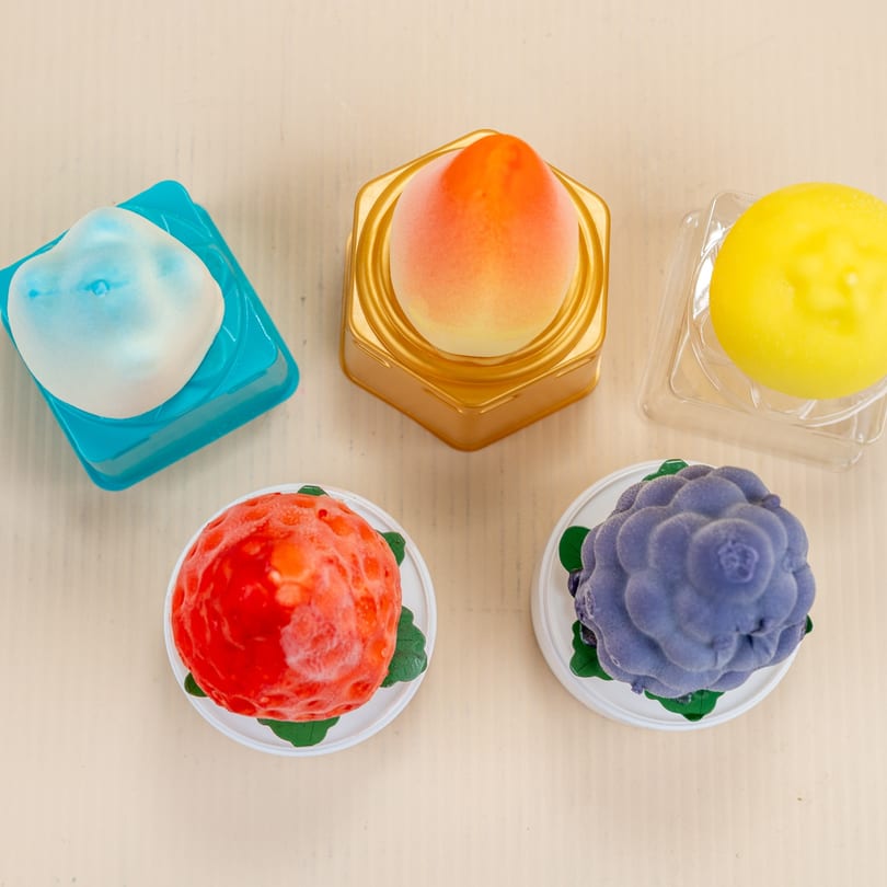 Five colorful fruit-shaped desserts, including peach, strawberry, blueberry, and lemon, on a light background.