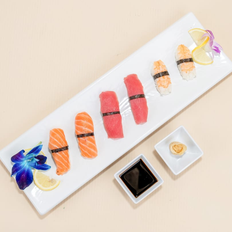 A white rectangular plate holds various sushi pieces, soy sauce, and ginger, garnished with orchids and lemon.