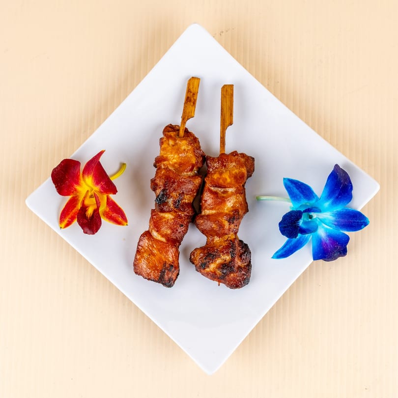 Two grilled chicken skewers on a white plate, garnished with red and blue orchids.