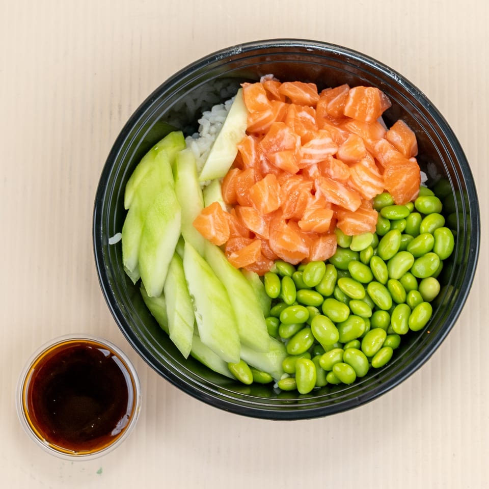 Litchi Ramen poke bowl with salmon, edamame, cucumber, and rice, with a side of sauce.