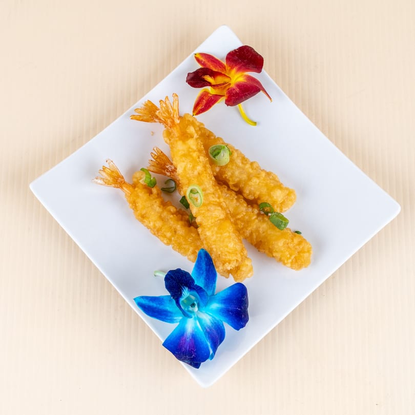 Golden fried shrimp tempura with green onions and two orchid garnishes on a white plate.