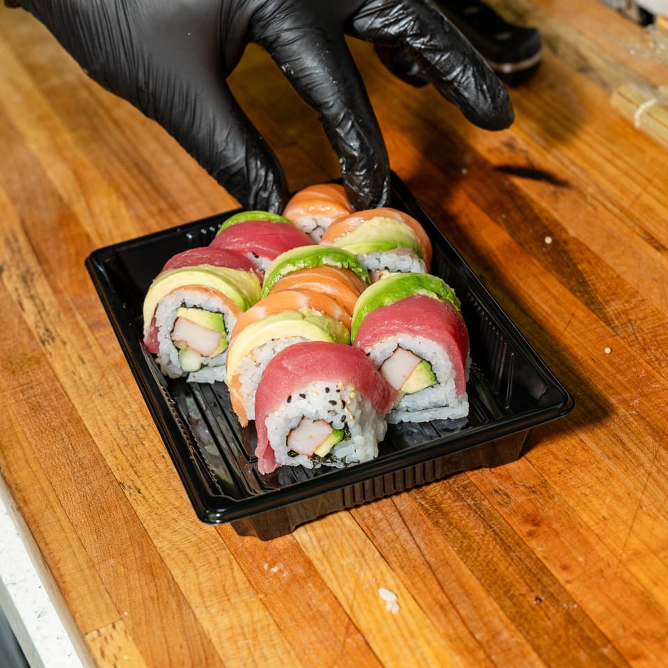 Gloved hands placing a black tray of colorful sushi rolls with avocado, salmon, and tuna on a wooden surface.