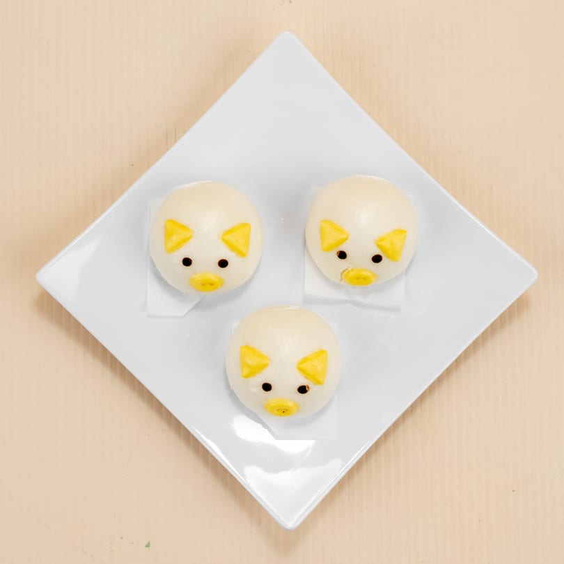 Three cute pig-shaped steamed buns with yellow ears and snouts on a white square plate.