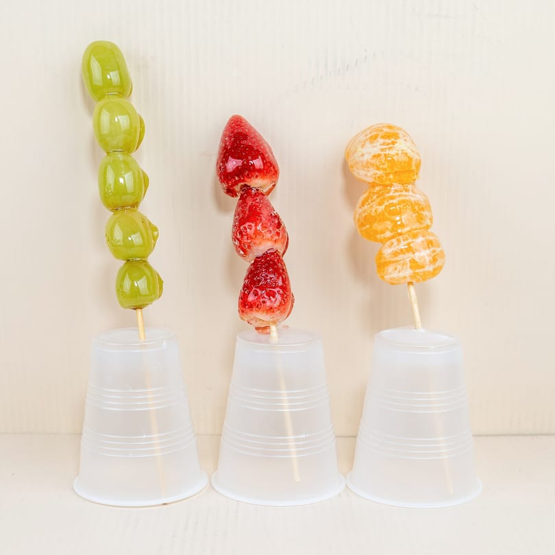 Three fruit skewers, with green grapes, strawberries, and mandarins, in clear plastic cups.